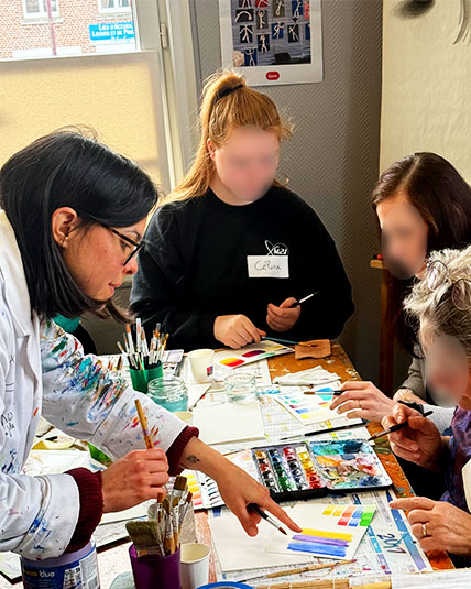 Julie Vigneron guide une élève pendant un atelier d’aquarelle, deux autres participantes peignent en arrière-plan