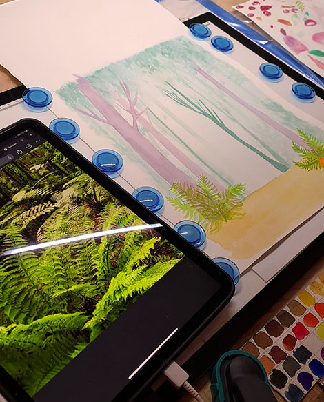 Table de travail de l’illustratrice avec une aquarelle en cours représentant la forêt de Marchiennes et une photo de fougère sur sa tablette en référence