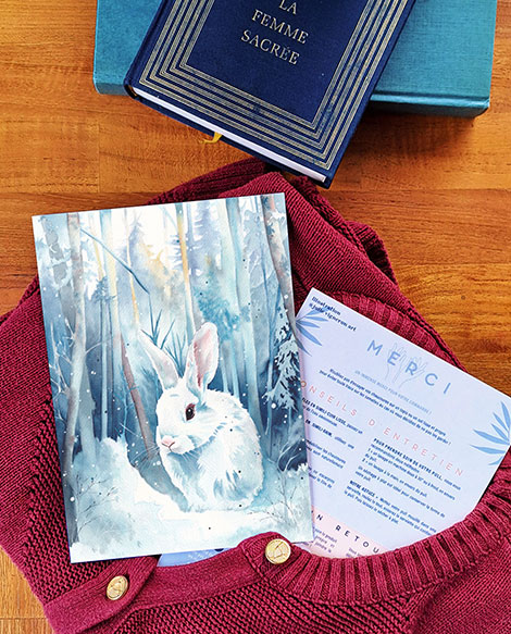 Carte de remerciement illustrée d'un lapin blanc dans un paysage enneigé par Julie Vigneron pour Minuit sur Terre, présentée avec un gilet bordeaux en laine recyclé de la marque et le verso de la carte imprimé avec remerciements et conseils d’entretien